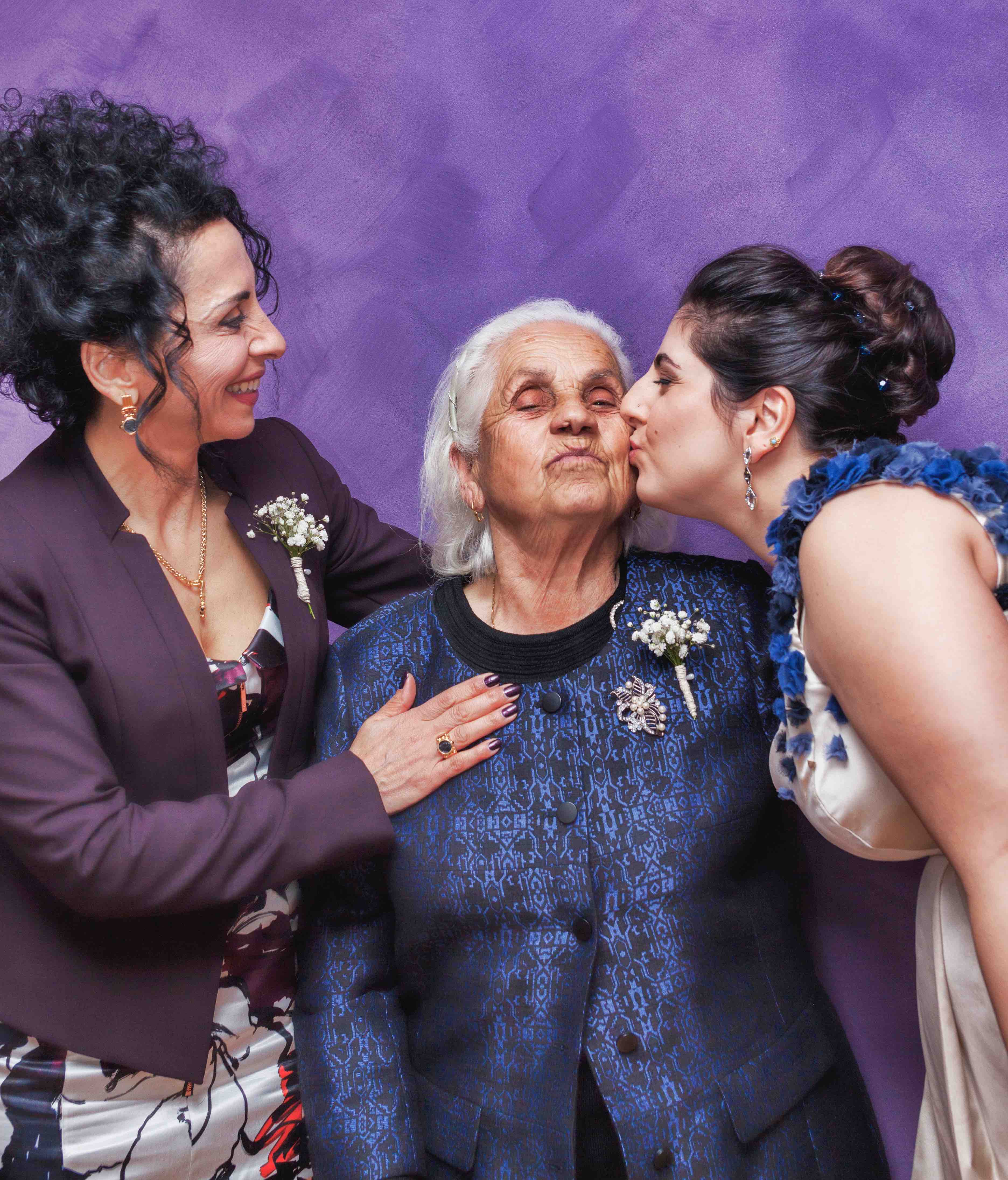 three women with one in purple and younger woman in white kissing older woman in blue
